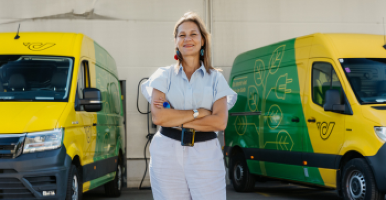 Eine Frau in heller Kleidung und langen blauen Haaren posiert mit verschränkten Armen zwischen zwei Kleintransportern, die gelb und grün gemustert sind und auf denen sich das Logo der österreichischen Post befindet.