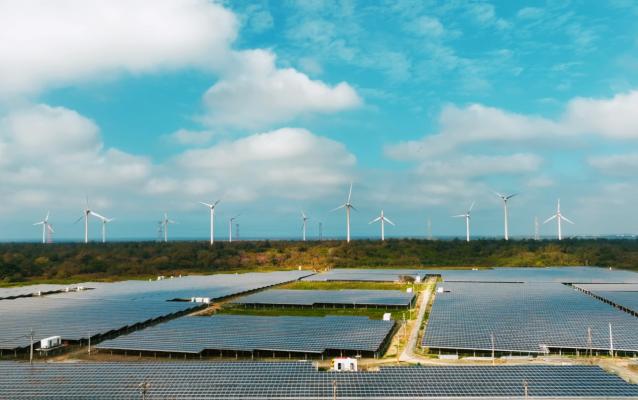 Luftaufnahme eines großen Solarparks mit zahlreichen Solarmodulen, die den Boden bedecken. Im Hintergrund ragen Windturbinen vor einem teilweise bewölkten blauen Himmel empor und demonstrieren eine Kombination erneuerbarer Energiequellen in einer grünen Landschaft.