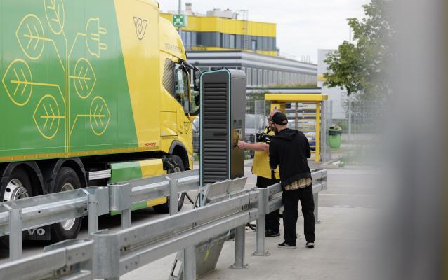 Zwei Arbeiter stehen neben einem großen gelb-grünen Lieferwagen mit umweltfreundlichen Blattmotiven in der Nähe einer Ladestation. Im Hintergrund sind ein modernes Industriegebäude und ein bewölkter Himmel zu sehen.