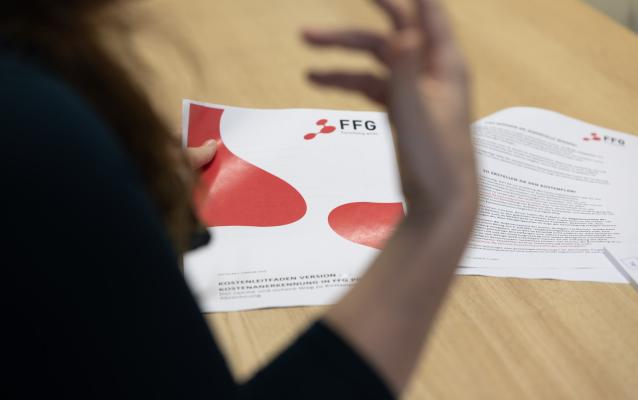 Eine Person hält ein Dokument mit dem FFG-Logo auf einem Tisch. Ihre Hand ist erhoben, möglicherweise gestikulierend. Das Dokument enthält gedruckten Text und rote Grafikelemente.