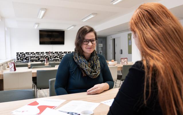 Zwei Personen sitzen an einem Tisch in einem hellen Raum mit Papieren, einer Kaffeetasse und einem Fernseher im Hintergrund. Eine Person trägt eine Brille und ein Kopftuch, während die andere lange rote Haare hat. Sie scheinen in ein Gespräch vertieft zu sein.